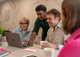 A man standing and demonstrating something on a laptop, watched by a man and a woman sitting either side.