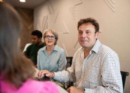 A man and a woman in an office sitting at a desk and looking at a woman sitting opposite whose face we can't see