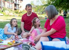 A family picnic, with a young girl speaking and her mother, father and daughter sitting listening to her
