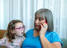 A mother sitting on a sofa and speaking into a phone, with her young daughter sitting next to her