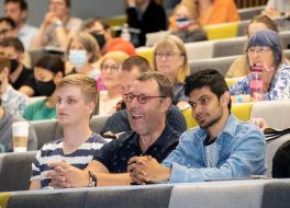 Three men sitting in a lecture theatre, looking to the left and laughing