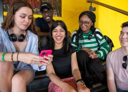 A group of five young people sitting on steps and looking at a phone that one of them is holding, and smiling