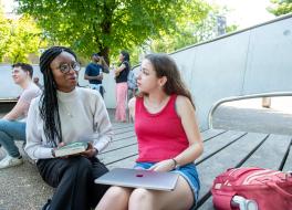 Two women seated in an outside setting and chatting, with other people in the background