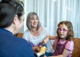 A young girl holding a toy and sitting with her mother while talking to a woman sitting opposite them