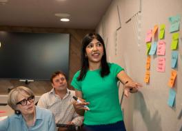 A woman standing beside a wall presenting to two people sitting beside her