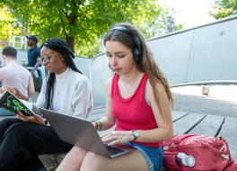 A woman wearing headphones and sitting outside typing on a laptop. Another woman reading a book is sitting next to her.
