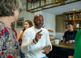 A man sitting in a coffee shop chatting to people standing around him and gesticulating with his hands