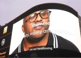 An advertising screen in Piccadilly Circus displaying a man looking at the camera, with the words 'Stamma' on it