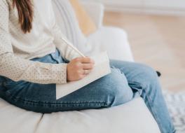 A woman sitting on a sofa writing in a diary. Her face is not in view