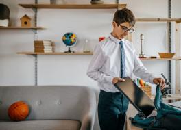A boy in a school uniform in a home setting, putting a laptop into a backpack
