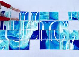 A piece of art consisting of coloured squares being put together by a person who is kneeling next to it