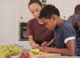 A boy cutting up a lime on a chopping board with his mother looking on. In the background is the father
