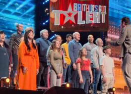 Members of a choir of mixed ages singing on a stage in front of a conductor. Above them is a logo saying 'Britain's Got Talent'