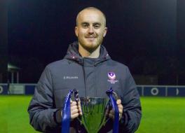 A man standing on a football pitch, holding a trophy and smiling