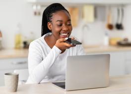 A woman sitting at a kitchen table in front of a laptop and speaking into a smartphone in her hand