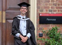 A young man in graduation robes, standing in front of a university door