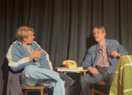 Two male actors, one younger and one older, sitting on a stage and talking in character.
