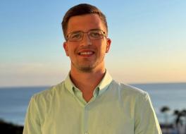 A young man smiling, with the ocean behind him