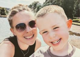 A woman wearing sunglasses standing next to her young son, in front of a lake. Both are smiling