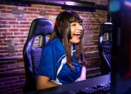 A woman at a desk wearing headphones, looking at a computer screen and laughing