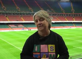 A woman standing in the seated area in a sporting stadium, with the pitch behind her