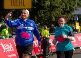 A man and woman running in a sponsored run. The man is holding a phone and taking a selfie