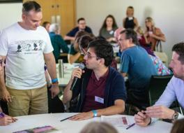 A man sitting at a table at a conference and talking into a microphone. Two men are sitting around him and a man is standing next to him, listening