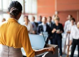 A woman standing a podium and speaking to an audience of people who are standing up