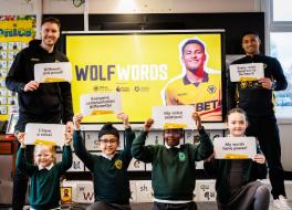 Two men standing either side of a screen, with four schoolchildren kneeling, all holding up paper with writing on it