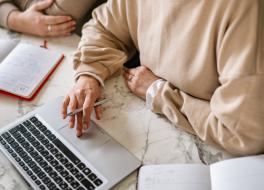 A typing on a laptop whilst holding a pen. Beside her is an other person. We can't see their faces.