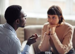 A man talking, with a woman listening to him, looking at him intently with her head resting on both hands