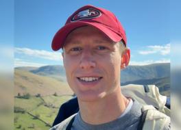 A man in an outdoor setting, wearing a baseball cap and smiling