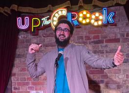 A man on a stage, smiling with his arms outstretched, in front of a microphone. The words 'Up the crook' are in lights behind him