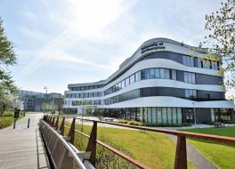 A modern university building surrounded trees and grass, with a bridge leading to it