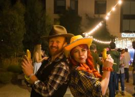 A man and a woman in cowboy outfits standing with their backs to each other and holding up bananas instead of guns