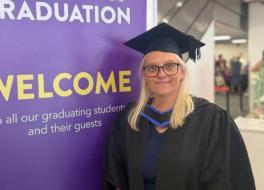 A woman in university gown and mortar board standing in front of a sign saying 'Welcome'