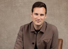 A man smiling, sitting in front of a brown background
