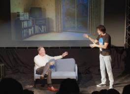Two male actors on a stage, one sitting on a sofa and the other standing