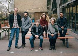 A group of people standing beside and sitting on a bench outdoors and looking at the camera
