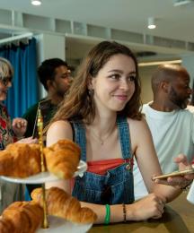 A young woman at a counter in a shop and smiling while using her phone to pay via a card reader held by somebody off screen.