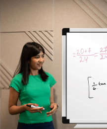 A woman holding a pen and standing beside a whiteboard which has writing on it