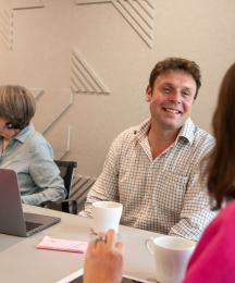 A man sitting at a desk chatting to a woman opposite whose face we can't see, with another woman sitting next to him chatting with a man next to her