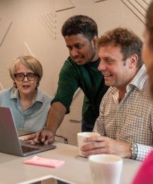 A man standing and demonstrating something on a laptop, watched by a man and a woman sitting either side.