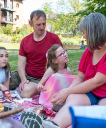 A family picnic, with a young girl speaking and her mother, father and daughter sitting listening to her