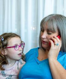 A mother sitting on a sofa and speaking into a phone, with her young daughter sitting next to her