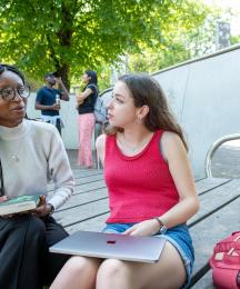 Two women seated in an outside setting and chatting, with other people in the background
