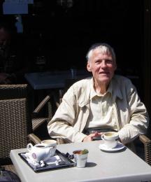 An elderly man sitting at a cafe table, looking at the camera and smiling