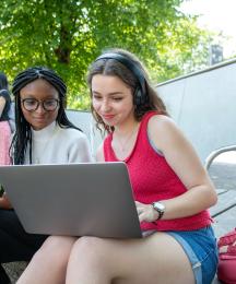 A woman in headphones showing something on her laptop to the woman sitting next to her.