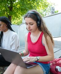 A woman wearing headphones and sitting outside typing on a laptop. Another woman reading a book is sitting next to her.