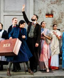 Seven actors on a stage, two of them waving, one carrying a suitcase and the others looking on 
